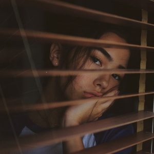 Eine Person stützt ihr Gesicht auf ihre Hand und blickt nachdenklich durch die teilweise geöffneten Jalousien, wobei weiches Licht und Schatten auf ihr Gesicht fallen.