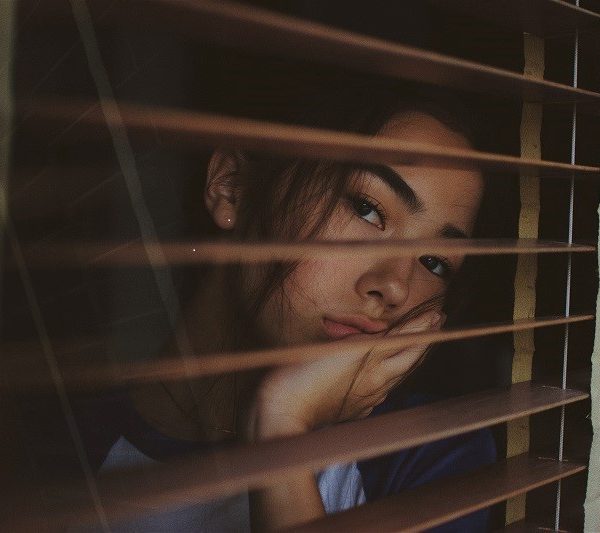 Eine Person stützt ihr Gesicht auf ihre Hand und blickt nachdenklich durch die teilweise geöffneten Jalousien, wobei weiches Licht und Schatten auf ihr Gesicht fallen.