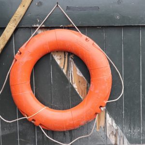 Ein orangefarbener Rettungsring hängt an einem Seil an einer verwitterten dunklen Holztür mit abblätternder Farbe und freiliegendem Holz.
