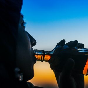 Eine Person in einer Kapuzenjacke trinkt bei Sonnenuntergang aus einer kleinen Glasflasche, wobei der Himmel im Hintergrund einen Farbverlauf von Blau und Orange zeigt.