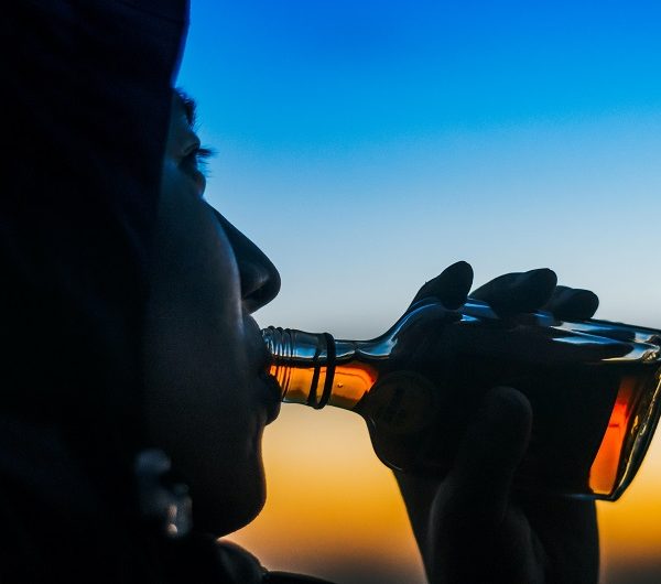 Eine Person in einer Kapuzenjacke trinkt bei Sonnenuntergang aus einer kleinen Glasflasche, wobei der Himmel im Hintergrund einen Farbverlauf von Blau und Orange zeigt.