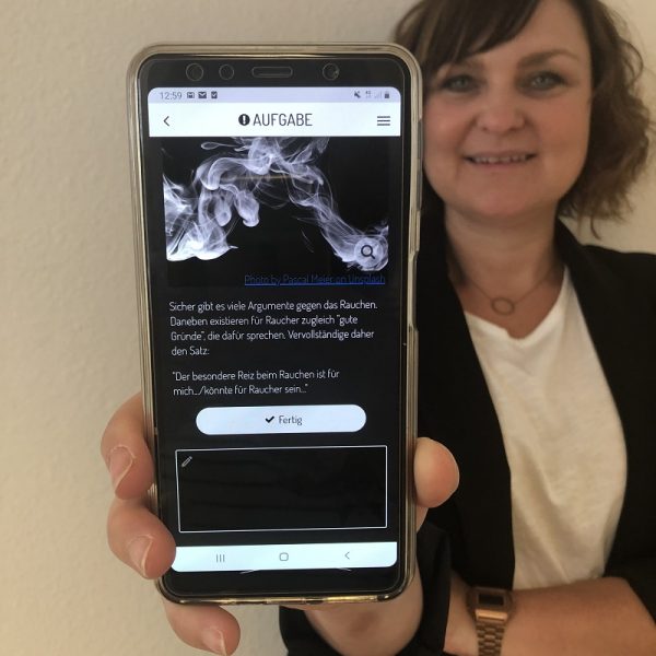 Eine Frau lächelt und hält ein Smartphone in die Kamera. Auf dem Bildschirm ist eine deutschsprachige Aufgabe zum Thema Rauchen zu sehen, mit Text und einem Bild von Rauch.