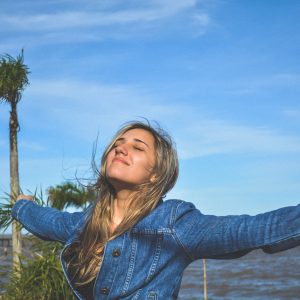 Eine Frau mit langem blondem Haar und Jeansjacke steht mit ausgestreckten Armen, geschlossenen Augen und einem Lächeln im Freien. Im Hintergrund sind Palmen, blauer Himmel und Wasser zu sehen.