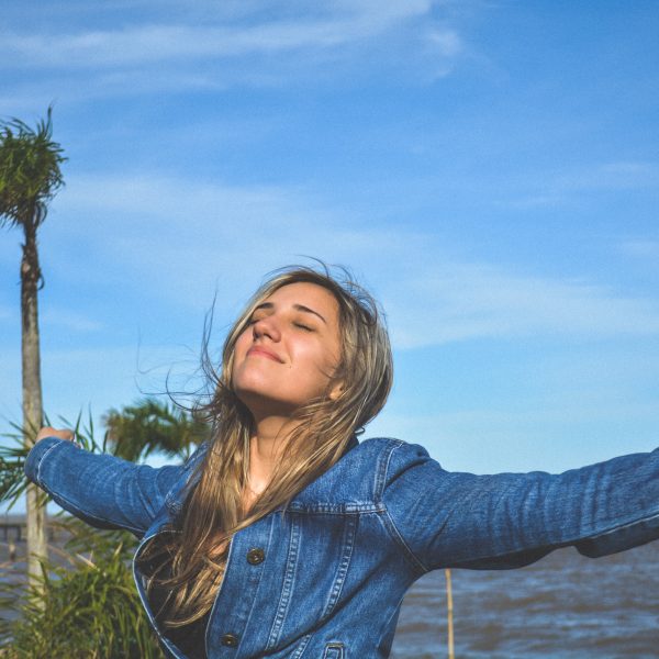 Eine Frau mit langem blondem Haar und Jeansjacke steht mit ausgestreckten Armen, geschlossenen Augen und einem Lächeln im Freien. Im Hintergrund sind Palmen, blauer Himmel und Wasser zu sehen.