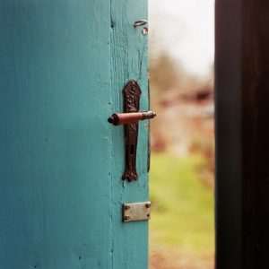 Eine Nahaufnahme einer alten blauen Holztür, die leicht angelehnt ist und einen alten Metallgriff und ein Schloss zeigt. Durch die Öffnung sind im Hintergrund unscharfe Grünpflanzen und eine Szene im Freien zu sehen.