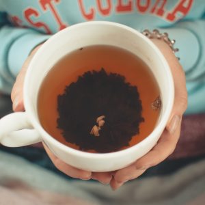 Eine Person, die ein hellblaues Sweatshirt trägt, hält eine große weiße Tasse, die mit Tee gefüllt ist. Ein dunkles Teeblattbüschel schwimmt in der Mitte des Getränks. Die Hände der Person sind sichtbar, und eine Decke bedeckt ihren Schoß.