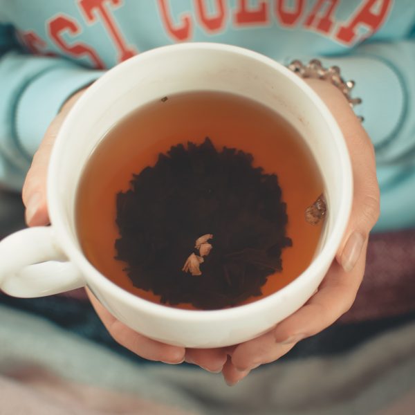 Eine Person, die ein hellblaues Sweatshirt trägt, hält eine große weiße Tasse, die mit Tee gefüllt ist. Ein dunkles Teeblattbüschel schwimmt in der Mitte des Getränks. Die Hände der Person sind sichtbar, und eine Decke bedeckt ihren Schoß.