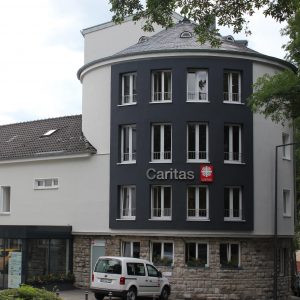 Ein modernes Gebäude mit dunklen und hellen Außenwänden, das über dem Eingang das "Caritas"-Logo trägt. Davor ist ein weißer Lieferwagen geparkt, und Bäume rahmen die Szene teilweise ein.