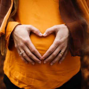 Eine Person in einem orangefarbenen Hemd formt mit ihren Händen eine Herzform über ihrem schwangeren Bauch und trägt zwei Ringe.