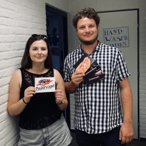 Eine Frau und ein Mann stehen in einem Haus und lächeln in die Kamera. Die Frau hält ein Feuervogel-Schild und der Mann ein Heft in der Hand. Hinter ihnen befindet sich eine Tür mit einem handgeschriebenen Hand-Wasch-Station-Schild.