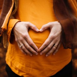 Eine Person, die ein orangefarbenes Oberteil trägt, formt mit ihren Händen über ihrem schwangeren Bauch eine Herzform, während sie im Freien steht, mit verschwommenem Herbstlaub im Hintergrund.