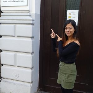 Eine Frau mit langen dunklen Haaren, die ein schwarzes Oberteil und einen grünen Rock trägt, lächelt und zeigt auf ein Schild mit der Aufschrift Suchthilfe Aachen Jugend- und Drogenberatung neben einer dunklen Holztür.