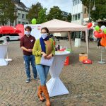 Zwei maskierte Personen stehen bei einer Standaktion im Freien an hohen Tischen mit Luftballons, Schildern und Infomaterialien. Im Hintergrund sind Gebäude und ein roter Lieferwagen zu sehen.