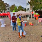 Zwei Personen mit Gesichtsmasken stehen bei einer Veranstaltung im Freien an einem Stehtisch. Auf einem gepflasterten Platz in einer Stadt sind Luftballons, Banner, ein Zeltdach und andere Veranstaltungsmaterialien aufgebaut. Im Hintergrund sind Bäume und Gebäude zu sehen.