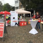 Ein Informationsstand für den Bundesweiten Aktionstag Glücksspielsucht mit Bannern, Tischen, Luftballons und zwei Personen, die in der Nähe stehen, eine mit einem Baguette in der Hand, die andere lächelt in die Kamera.