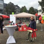 Drei Personen stehen an einem Informationsstand im Freien mit Tischen, Bannern, Luftballons und Werbeartikeln auf einem Platz mit Kopfsteinpflaster. Im Hintergrund sind Gebäude, Bäume und der bewölkte Himmel zu sehen.