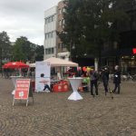 Ein Straßenstand im Freien, der mit einem roten Schild, einem Infotisch und Luftballons für den Aktionstag wirbt. Menschen sind versammelt; ein Kamerateam filmt. Der WOOLWORTH-Laden und andere Geschäfte sind im Hintergrund zu sehen.