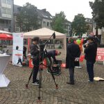 Menschen bauen ihre Ausrüstung unter einem Vordach auf einem gepflasterten Stadtplatz auf. Eine Kamera auf einem Stativ ist auf sie gerichtet, während andere Ballons und Schilder halten. Gebäude und Bäume säumen den Hintergrund an einem bewölkten Tag.