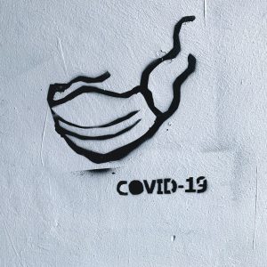 Eine schwarze Schablonenzeichnung einer Gesichtsmaske und der Text "COVID-19" sind auf eine strukturierte weiße Wand gesprüht.