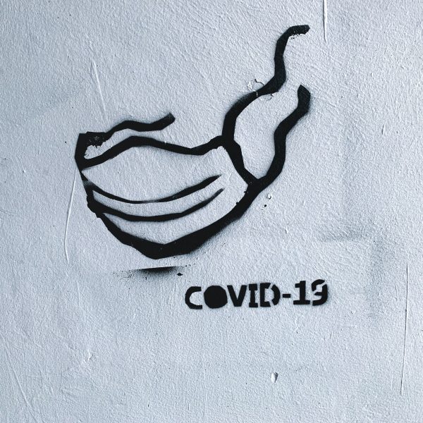 Eine schwarze Schablonenzeichnung einer Gesichtsmaske und der Text "COVID-19" sind auf eine strukturierte weiße Wand gesprüht.