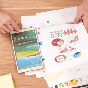Eine Person betrachtet bunte Diagramme und Grafiken auf bedruckten Papieren an einem Schreibtisch, daneben ein Tablet, Haftnotizen, Stifte und Textmarker. Der Bildschirm des Tablets ist eingeschaltet und steht auf dem Kopf.