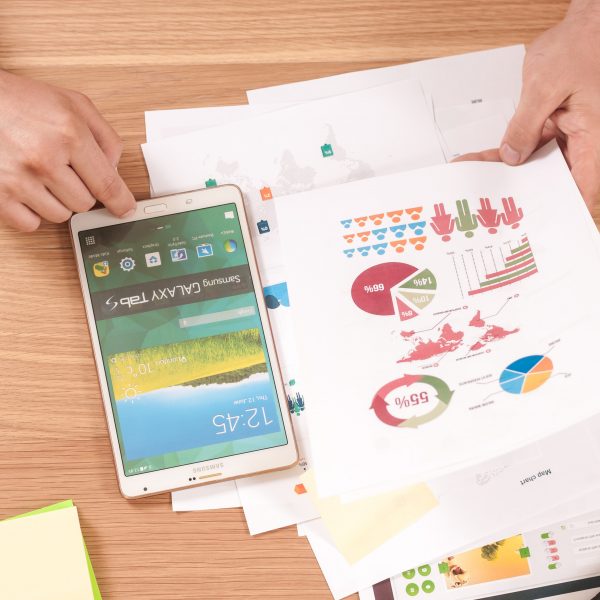 Eine Person betrachtet bunte Diagramme und Grafiken auf bedruckten Papieren an einem Schreibtisch, daneben ein Tablet, Haftnotizen, Stifte und Textmarker. Der Bildschirm des Tablets ist eingeschaltet und steht auf dem Kopf.