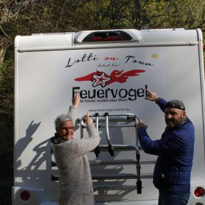 Zwei lächelnde Menschen stehen am Heck eines weißen Wohnmobils und zeigen auf ein großes Feuervogel-Logo und den Text Lotti on Tour. Im Hintergrund sind Bäume mit Herbstlaub zu sehen.