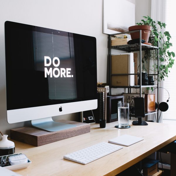 Ein aufgeräumtes Heimbüro mit einem großen iMac, auf dessen Bildschirm der Schriftzug DO MORE. zu lesen ist, einer Tastatur, einem Glas Wasser, Pflanzen, Büchern und verschiedenen Schreibtisch-Accessoires neben einem Fenster, das natürliches Licht hereinlässt.