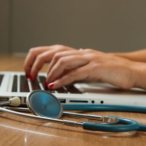 Eine Person mit rot lackierten Fingernägeln tippt auf einem Laptop, während ein Stethoskop auf dem hölzernen Schreibtisch neben ihr liegt.
