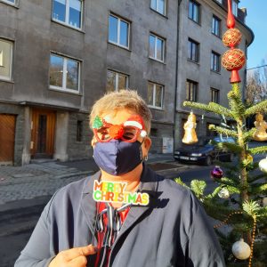 Eine Person, die eine festliche Weihnachtsbrille und eine Gesichtsmaske trägt und ein "Frohe Weihnachten"-Schild in der Hand hält, steht neben einem geschmückten Weihnachtsbaum in einer sonnigen Straße.