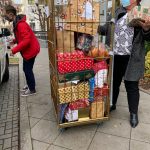 Zwei maskierte Personen laden einen großen Metallwagen mit eingepackten Weihnachtsgeschenken auf einen Bürgersteig. Eine Person hält ein Geschenk in der Hand, während die andere in der Nähe eines am Straßenrand geparkten Autos steht.