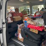 Ein Lieferwagen mit offener Seitentür, vollgepackt mit zahlreichen bunt eingepackten Geschenken und Geschenktüten, teilweise mit Anhängern, die sich auf den Sitzen und dem Boden stapeln.