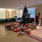 Ein geschmückter Weihnachtsbaum steht in der Mitte eines Raumes, umgeben von zahlreichen eingepackten Geschenken. Der Raum ist mit modernen Möbeln, Weihnachtspostern an den Wänden und großen Fenstern ausgestattet, die natürliches Licht hereinlassen.
