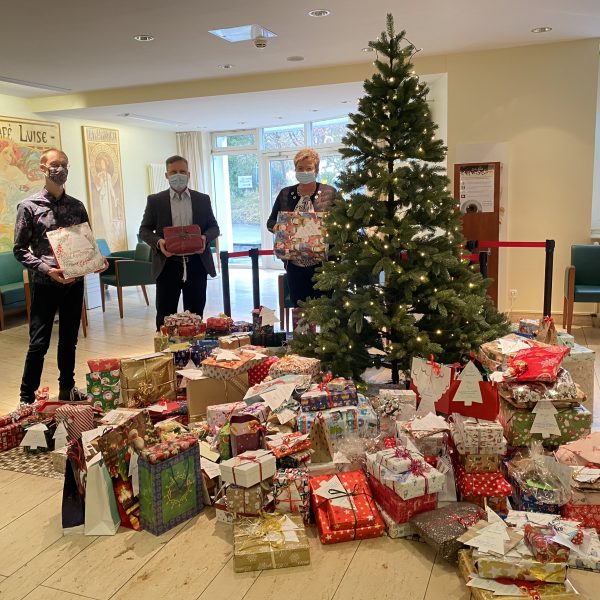 Drei maskierte Personen stehen neben einem geschmückten Weihnachtsbaum, unter dem sich viele eingepackte Geschenke stapeln. Der Raum ist hell mit festlichen Dekorationen und Gemälden an den Wänden.