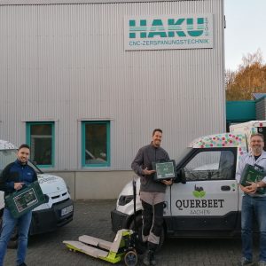 Drei Männer stehen lächelnd vor zwei gebrandeten Lieferwagen und einem Gebäude mit der Aufschrift HAKU GmbH CNC-ZERSPANUNGSTECHNIK. Jeder von ihnen hält eine grüne Kiste in der Hand, ein gelber Hubwagen steht vor ihnen. Die Umgebung ist industriell geprägt.