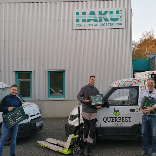 Drei Männer stehen lächelnd vor zwei gebrandeten Lieferwagen und einem Gebäude mit der Aufschrift HAKU GmbH CNC-ZERSPANUNGSTECHNIK. Jeder von ihnen hält eine grüne Kiste in der Hand, ein gelber Hubwagen steht vor ihnen. Die Umgebung ist industriell geprägt.