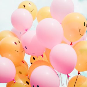 Ein Haufen gelber und rosafarbener Luftballons, einige mit einfachen Smiley-Gesichtern bemalt, schweben vor einem strahlend blauen Himmel mit vereinzelten Wolken.