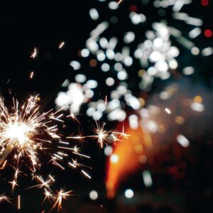 Eine Nahaufnahme einer brennenden Wunderkerze mit hellen, funkelnden Spuren vor einem dunklen Hintergrund, mit verschwommenem Feuerwerk oder Lichtern, die in der Ferne leuchten.