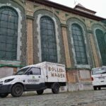 Zwei weiße Lieferwagen mit dem "Querbeet Aachen"-Logo stehen auf dem Kopfsteinpflaster vor einem großen Backsteingebäude mit hohen Rundbogenfenstern. Ein Lieferwagen trägt die Aufschrift SÜLLER-WAGEN auf seiner Seite.