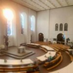 Ein geräumiger, moderner Kirchenraum mit hohen Decken, großen Bogenfenstern, die das Sonnenlicht hereinlassen, geschwungenen Holzbänken und zeitgenössischen Skulpturen, die den Raum schmücken.