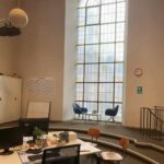 Modernes Büro mit großem Bogenfenster, zwei blauen Stühlen und einem kleinen Tisch am Fenster, Schreibtischen und Stühlen im Vordergrund, Whiteboard, Wandkalender und einer Uhr an der Wand. Der Raum ist von natürlichem Licht durchflutet.