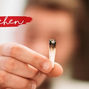 Eine Person hält einen teilweise verbrannten Joint in die Kamera, auf dem in weißer Schreibschrift auf einem roten Farbstreifen in der oberen linken Ecke das Wort verstehen geschrieben steht. Der Hintergrund ist unscharf.