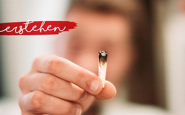 Eine Person hält einen teilweise verbrannten Joint in die Kamera, auf dem in weißer Schreibschrift auf einem roten Farbstreifen in der oberen linken Ecke das Wort verstehen geschrieben steht. Der Hintergrund ist unscharf.