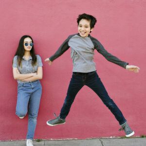 Ein Mädchen mit Sonnenbrille steht mit verschränkten Armen an einer rosafarbenen Wand, während ein lächelnder Junge energisch neben ihr herumspringt, beide in lässigen Jeans und Tops.