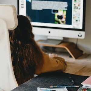 Eine Person mit langen Haaren sitzt an einem Schreibtisch und arbeitet an einem Computer mit einem großen Monitor. Auf dem Bildschirm werden Texte und Bilder angezeigt. Auf dem Schreibtisch im Vordergrund liegen Büromaterial, Notizbücher und Stifte verstreut.