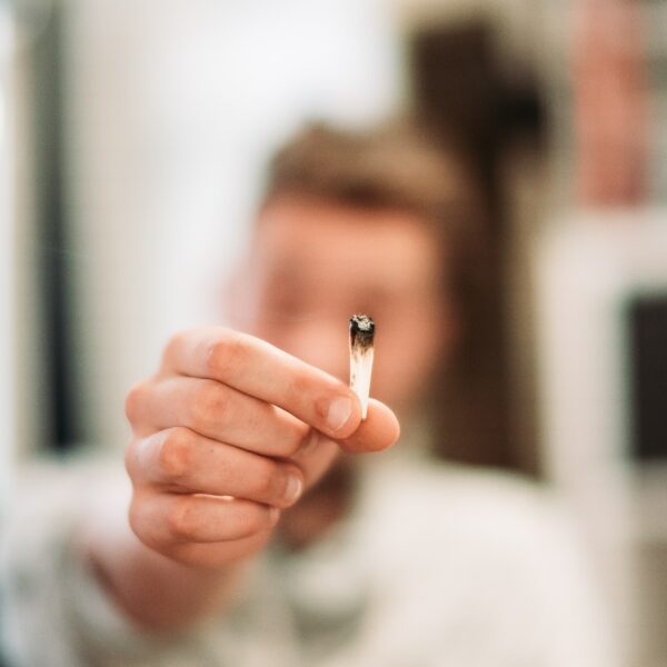 Eine Person, die einen angezündeten, teilweise gerauchten Joint zwischen den Fingern hält, wobei der Fokus auf der Hand und dem Joint liegt, während das Gesicht der Person im Hintergrund verschwommen ist.