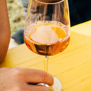 Eine Person, die einen goldenen Ring trägt, hält ein Glas Roséwein am Stiel über einem hellgelben Holztisch. Eine weitere Person ist unscharf im Hintergrund zu erkennen.