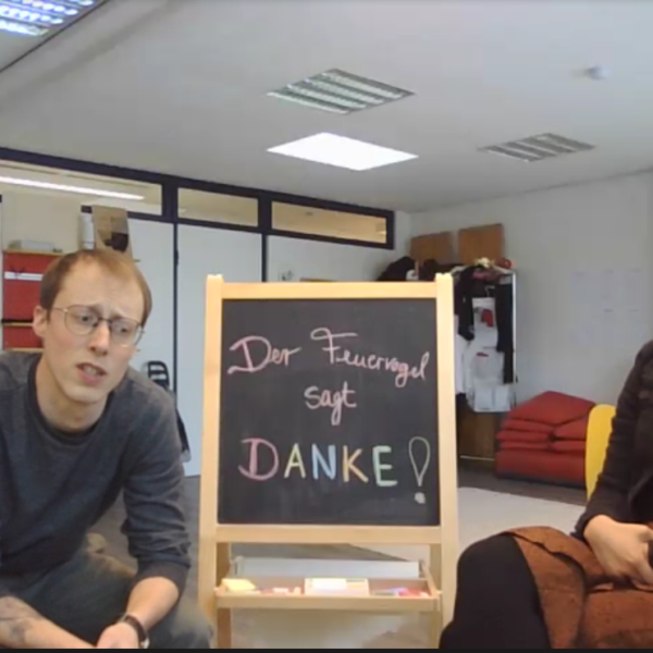 Zwei Personen sitzen in einem Raum neben einer kleinen Kreidetafel, auf der auf Deutsch Der Feuervogel sagt DANKE! steht. Der Raum ist mit Büromöbeln, einem Kleiderständer und einer roten Couch im Hintergrund ausgestattet.
