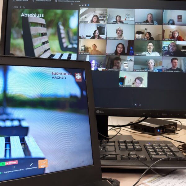 Ein Laptop und ein Desktop-Monitor zeigen ein virtuelles Meeting mit mehreren Teilnehmern auf dem Bildschirm; der Laptop zeigt außerdem ein verschwommenes Bild von Bänken und das Logo der Suchthilfe Aachen. Auf dem Schreibtisch befinden sich eine Tastatur und eine Maus.