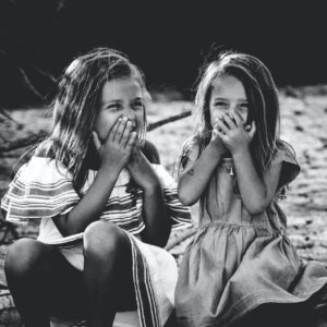 Zwei junge Mädchen sitzen im Freien auf einem umgestürzten Baum, lächeln und halten sich den Mund mit den Händen zu, als würden sie lachen. Das Foto ist in schwarz-weiß gehalten, mit einem natürlichen, sandigen Hintergrund.
